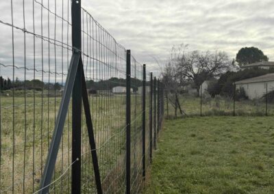 Clôture de jardin en grillage soudé noir fixée sur des poteaux en T avec jambes de force, bordant un terrain herbeux sous un ciel nuageux
