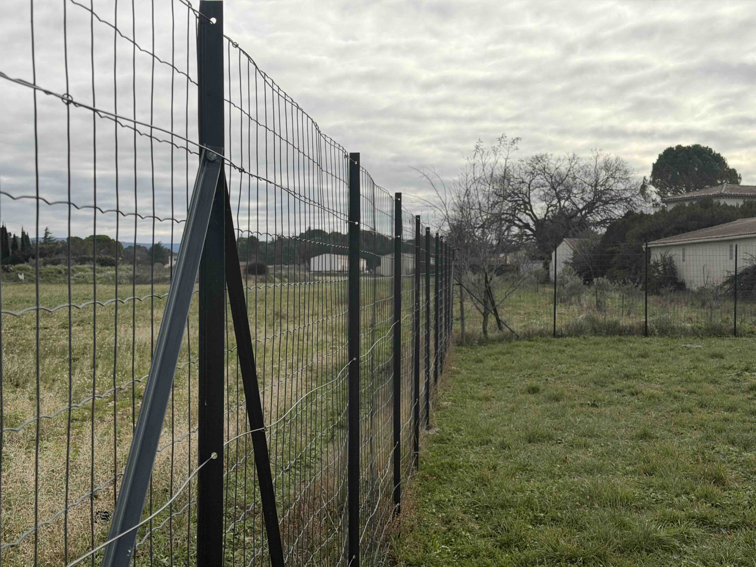 Clôture de jardin en grillage soudé noir fixée sur des poteaux en T avec jambes de force, bordant un terrain herbeux sous un ciel nuageux